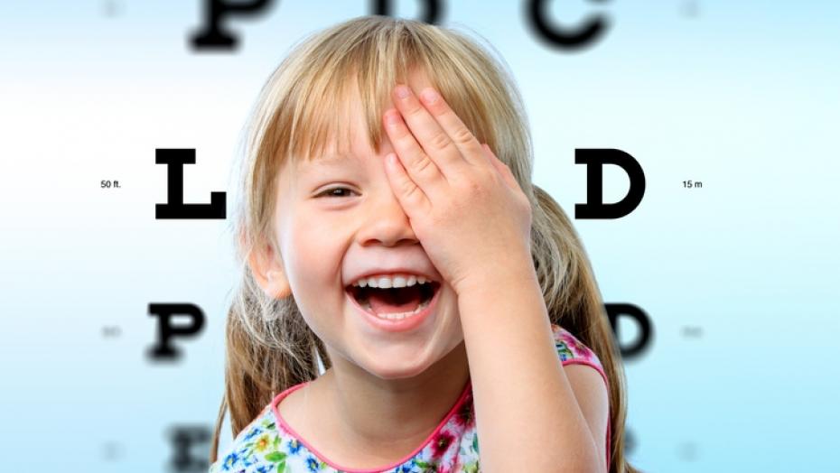 Young female smiling and using her hand to cover her left eye, she is in front of a background that resembles a Snellen eye chart